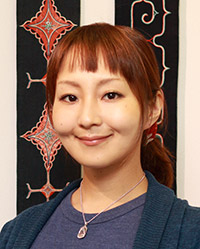 顔写真：アイヌ文様のタペストリーを背景に笑顔の三橋とらさん